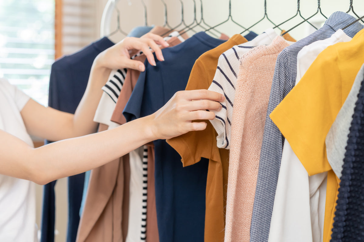 A person's hands browsing through a rack of colorful clothing, including navy blue, mustard yellow, pink, and white striped garments.