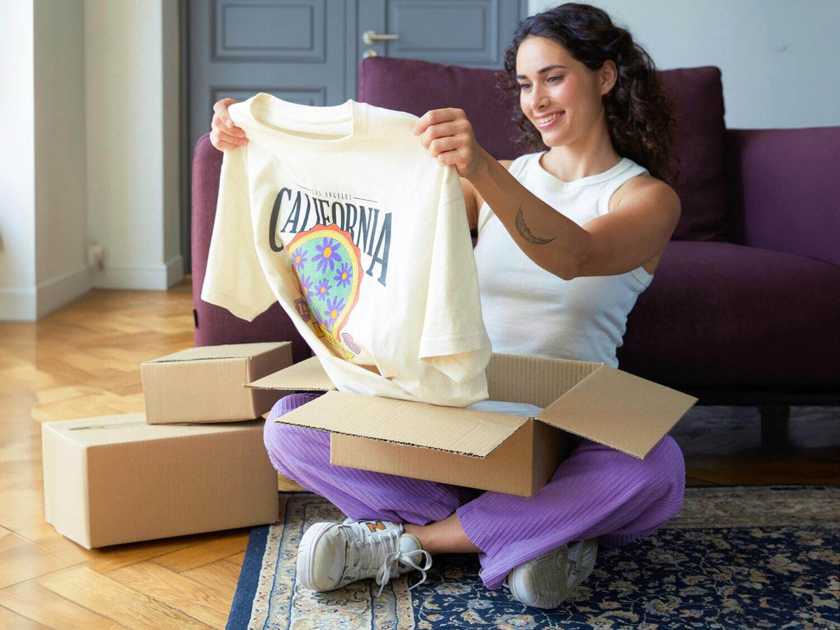 A woman sitting on a patterned rug in a living room holds up a cream-colored t-shirt with a colorful 'Cali' graphic. Several cardboard boxes are stacked around her, with a purple couch in the background.